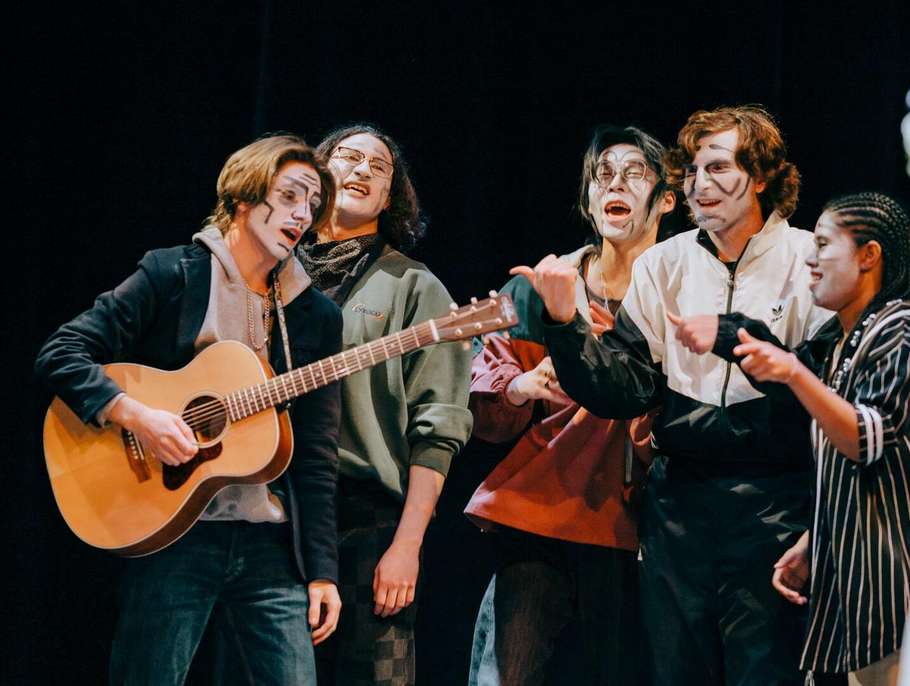 Group of student actors wearing theatrical face paint performing a musical scene with an acoustic guitar on stage.